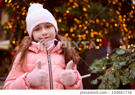 Cute little girl having fun at Christmas fair, showing thumbs up at camera, standing by illuminated Christmas tree 109661736