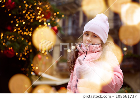 Photography with bokeh of authentic little child girl standing by illuminated Christmas tree outdoors at Christmas fair. 109661737