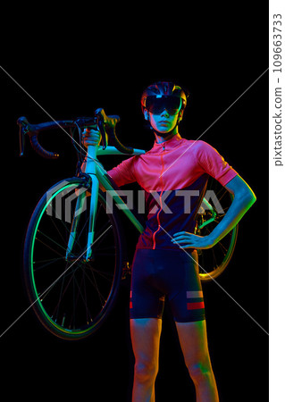 Young man in sportswear, helmet and googles holding bicycle on shoulder isolated over black studio background in neon light 109663733