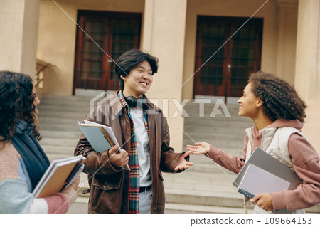 Cheerful students talking each other standing near university campus and holding books and laptop Cheerful students talking each other standing near university campus and holding books and laptop 109664153