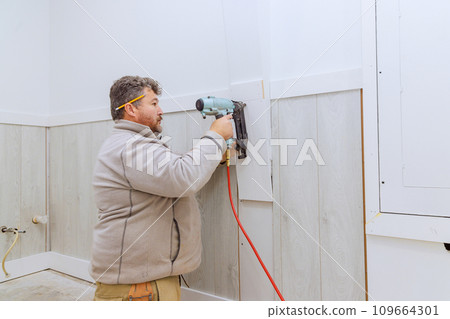 There is worker from contractor nailing wooden trim moldings on walls of room using air gun 109664301