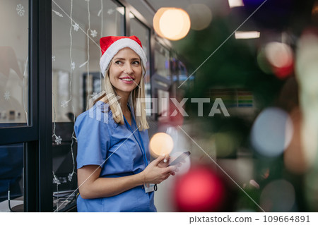 Portrait of beautiful nurse with christmas hat in hospital corridor, holding smartphone. Working in hospital on Christmas day, Christmas Eve. Female doctor working a Christmas shift and can't be with Portrait of beautiful nurse with christmas hat in hospital corridor, holding smartphone. Working in hospital on Christmas day, Christmas Eve. Female doctor working a Christmas shift and can't be with 109664891
