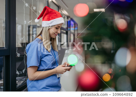Portrait of beautiful nurse with christmas hat in hospital corridor, holding smartphone. Working in hospital on Christmas day, Christmas Eve. Female doctor working a Christmas shift and can't be with 109664892