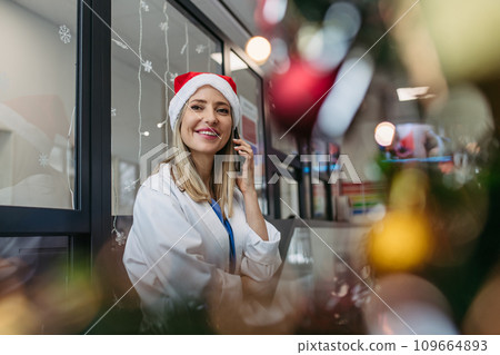 Portrait of beautiful doctor with christmas hat making call. Working in hospital on Christmas day, Christmas Eve. Female doctor working a Christmas shift and can't be with her family. 109664893
