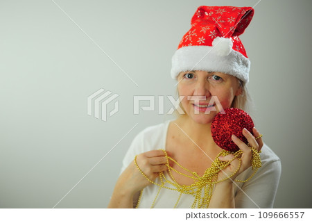 An adult woman on a white background stands smiling at her cap hat Christmas Christmas tree toys bright red New year party invitation Nice beautiful woman happy new year greetings 109666557