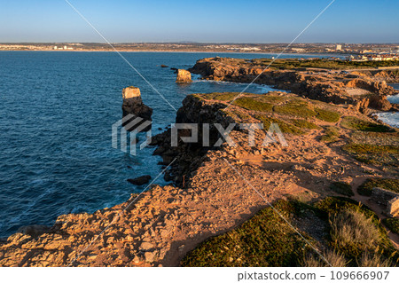 Bird's-eye view of ocean waves break on rocky shores pf Peniche, Portugal. Summer sunset haze, little foliage and rocky cliffs, peninsula and rocks, fishing town, horizon.  109666907