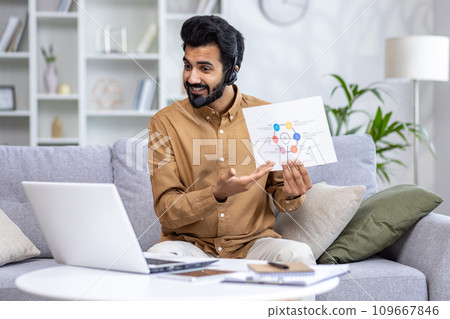 Smiling Indian male businessman, teacher, trainer showing documents and talking online via laptop. Sitting at home on the sofa in a headset. Smiling Indian male businessman, teacher, trainer showing documents and talking online via laptop. Sitting at home on the sofa in a headset. 109667846