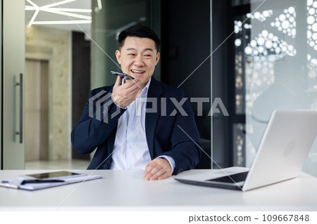 Asian young man businessman sitting in the office at the desk with a laptop, and talking on the mobile phone through the loudspeaker. Asian young man businessman sitting in the office at the desk with a laptop, and talking on the mobile phone through the loudspeaker. 109667848