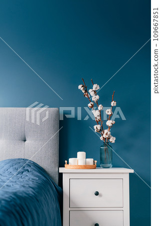 Stylish modern bedroom in dark colors. Cozy interior with navy blue walls, home decor. Bed with gray fabric headboard, blue blanket, bedside table, vase with natural cotton flowers, candles on tray 109667851