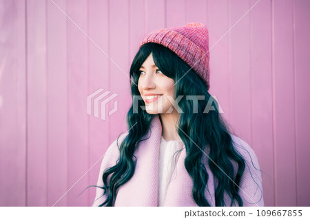 Stylish young smiling hipster woman with color hair wearing pink coat and knitted hat on pink wooden background. Urban seasonal city street fashion. Barbiecore style. Selective focus. Stylish young smiling hipster woman with color hair wearing pink coat and knitted hat on pink wooden background. Urban seasonal city street fashion. Barbiecore style. Selective focus. 109667855