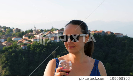 Young cheerful woman in sunglasses drinking red at sunset, beautiful European town on the background 109668593