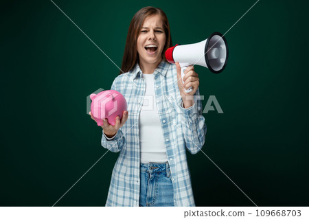 A cute young woman in a plaid shirt talks about financial literacy while holding a piggy bank in her 109668703