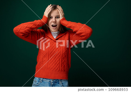 Surprised woman with brown hair dressed in a red sweater and jeans holds her head on a green Surprised woman with brown hair dressed in a red sweater and jeans holds her head on a green 109668933