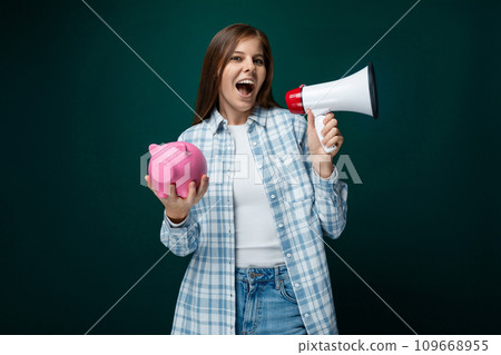 Young European woman dressed in a plaid shirt and jeans holds a piggy bank and talks about 109668955