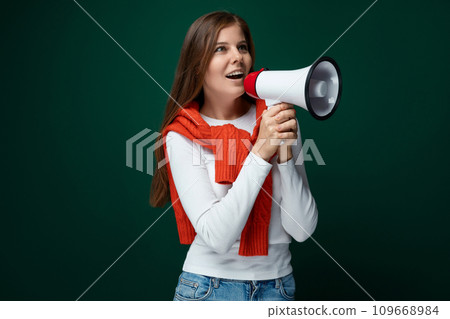 Positive young woman tied a red sweater over her shoulders and speaks into a loudspeaker Positive young woman tied a red sweater over her shoulders and speaks into a loudspeaker 109668984