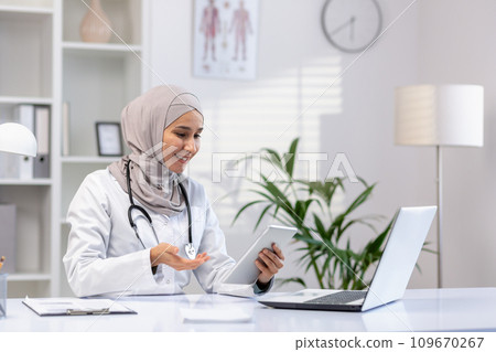 Arab Muslim woman doctor sits at a table in the office in front of a laptop, conducts an online consultation, an appointment with a patient, holds a tablet in her hands. Arab Muslim woman doctor sits at a table in the office in front of a laptop, conducts an online consultation, an appointment with a patient, holds a tablet in her hands. 109670267