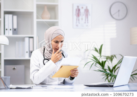 Upset arab female doctor in hijab, holding an envelope, sitting at the table in her office, bad test results, unpleasant news. Upset arab female doctor in hijab, holding an envelope, sitting at the table in her office, bad test results, unpleasant news. 109670273