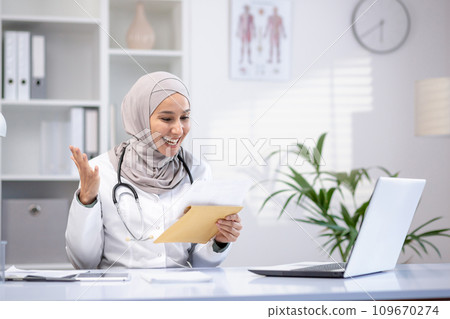 Happy female doctor in hijab holding letter in hands, sitting at table in hospital office, happy with good news. 109670274