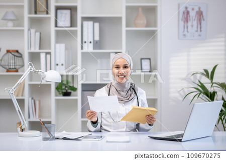 Portrait of a happy Muslim Arab female doctor sitting at a table in an office in a private clinic, holding an envelope, good news, good results, smiling, looking at the camera. Portrait of a happy Muslim Arab female doctor sitting at a table in an office in a private clinic, holding an envelope, good news, good results, smiling, looking at the camera. 109670275
