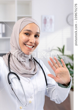 A Muslim female doctor in a hijab conducts an online appointment with patients on the phone, waves to the camera, view from the phone. A Muslim female doctor in a hijab conducts an online appointment with patients on the phone, waves to the camera, view from the phone. 109670276