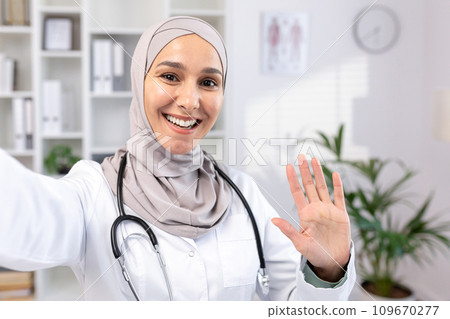 Beautiful asian muslim female doctor in hijab and suit taking selfie photo while resting. A doctor makes a video call, smiling and waving as he greets his family from his workplace Beautiful asian muslim female doctor in hijab and suit taking selfie photo while resting. A doctor makes a video call, smiling and waving as he greets his family from his workplace 109670277