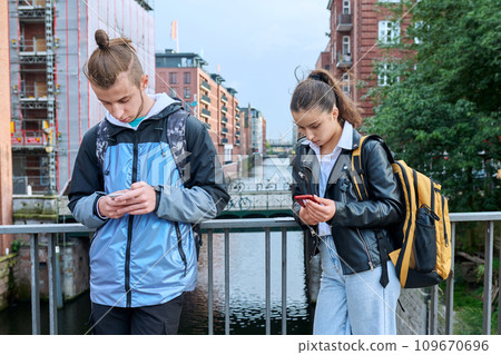 Teenage friends guy and girl standing together using smartphones Teenage friends guy and girl standing together using smartphones 109670696