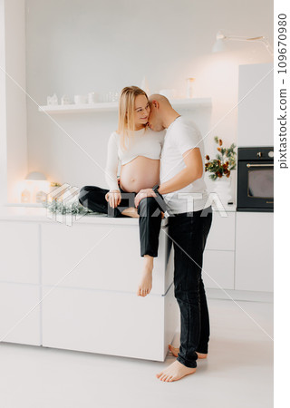 Couple seems very happy. A handsome husband is embracing his beautiful pregnant wife next to the window in the living room. 109670980
