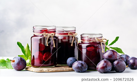 Ripe plums and plum compote in glass jars on the table. Home canned food and harvest of appetizing plums on a white background. 109672026