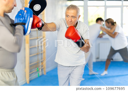 Coach and elderly man pupil are engaged during training, boxing classes. Male trainer holds makiwara and helps student to work out force of blow, to box quickly and effectively Coach and elderly man pupil are engaged during training, boxing classes. Male trainer holds makiwara and helps student to work out force of blow, to box quickly and effectively 109677695