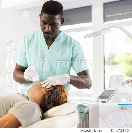 African male doctor uses scriber beauty machine nozzle for gentle skin peeling for man client African male doctor uses scriber beauty machine nozzle for gentle skin peeling for man client 109678480
