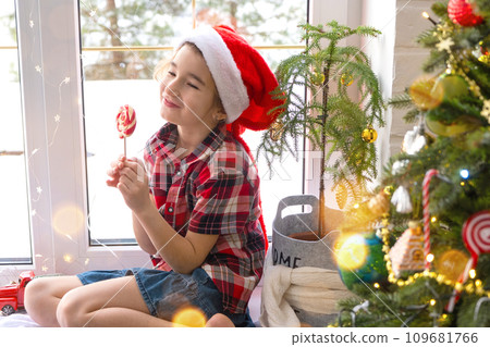 Happy Girl in a Santa hat sits on the windowsill of a house near the Christmas tree and puts sweet round caramel on a stick. Child is having fun and making faces, waiting for Christmas and New year 109681766