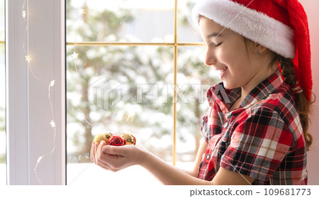 Girl in a Santa hat sits on the windowsill of a house near the Christmas tree and holding glass balls in his hands. Child is having fun, believes in a miracle and waiting for Christmas and New year 109681773