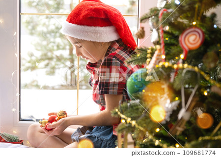 Girl in a Santa hat sits on the windowsill of a house near the Christmas tree and holding glass balls in his hands. Child is having fun, believes in a miracle and waiting for Christmas and New year 109681774