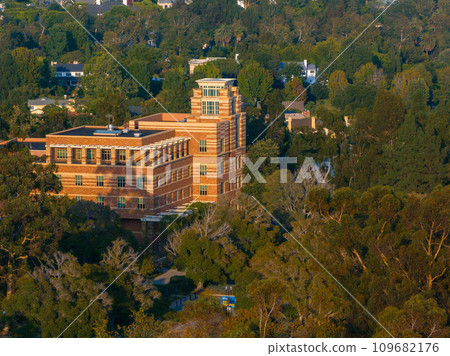 Aerial view of a modern multi-story building amidst a tranquil suburban landscape bathed in the warm, golden light of a late afternoon sun, surrounded by lush greenery. Aerial view of a modern multi-story building amidst a tranquil suburban landscape bathed in the warm, golden light of a late afternoon sun, surrounded by lush greenery. 109682176