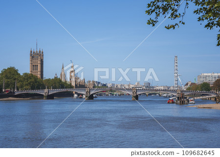 Big Ben and Millennium Wheel at banks of river. Westminster bridge over Thames water with blue sky in background. Scenic view of famous tourist attraction in city during sunny day. 109682645