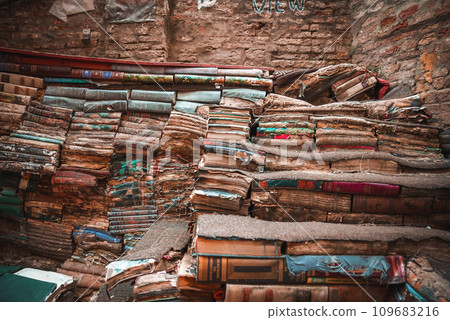 A dimly lit room in an old, weathered building in Venice. Sunlight streams in, casting a warm glow on a haphazard pile of old books, creating a cozy and inviting space. 109683216