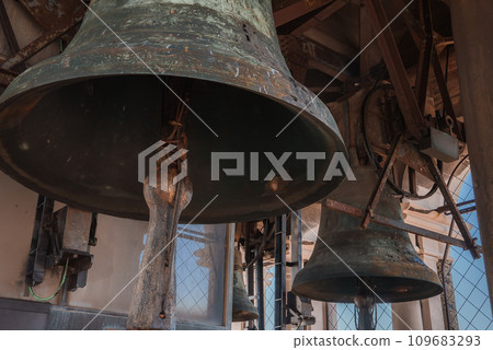 Three bells hang from the ceiling of a dimly lit church in Venice, Italy, creating a serene and peaceful atmosphere. The traditional Venetian architecture adds to the sense of tranquility. Three bells hang from the ceiling of a dimly lit church in Venice, Italy, creating a serene and peaceful atmosphere. The traditional Venetian architecture adds to the sense of tranquility. 109683293