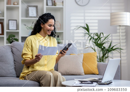 Happy hispanic woman working in home office sitting on sofa in front of laptop, holding phone and documents in hands, remote work. Happy hispanic woman working in home office sitting on sofa in front of laptop, holding phone and documents in hands, remote work. 109683350