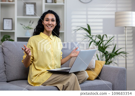 A smiling girl with a laptop on her lap sits on the sofa in the living room, spreads her arms and looks at the camera. A smiling girl with a laptop on her lap sits on the sofa in the living room, spreads her arms and looks at the camera. 109683354