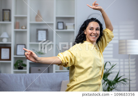Cheerful young girl dancing at home in the living room near the sofa, lifestyle, cheerful hispanic woman, carefree and relaxing. Cheerful young girl dancing at home in the living room near the sofa, lifestyle, cheerful hispanic woman, carefree and relaxing. 109683356