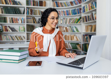 Smiling Indian businesswoman in headset sits at laptop in room, communicates with clients online, works in call center, looks at screen. Smiling Indian businesswoman in headset sits at laptop in room, communicates with clients online, works in call center, looks at screen. 109683397