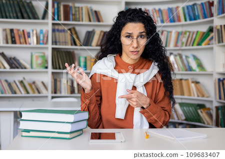 Dissatisfied hispanic woman teacher conducts online lesson sitting at desk in office, explains unclear topic, view from webcam. Dissatisfied hispanic woman teacher conducts online lesson sitting at desk in office, explains unclear topic, view from webcam. 109683407