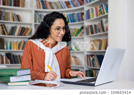 Satisfied female student listens to an online lecture on a laptop, writes something in a notebook, sits at a table in an office, distance learning. 109683414