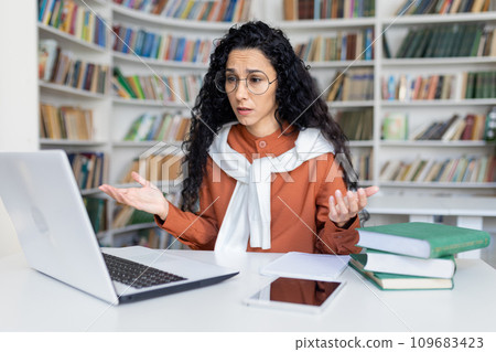 Upset female worker sitting at table in front of laptop, spreading hands, making video call, sad because of faulty equipment. 109683423