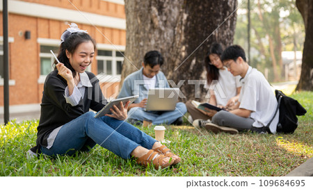 A positive Asian female college student is using her tablet while sitting with her friends in a park 109684695