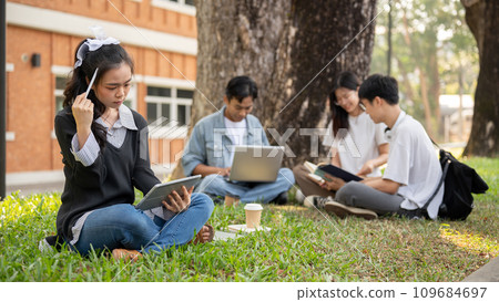 A focused Asian female college student is using her tablet while sitting in a park with her friends 109684697