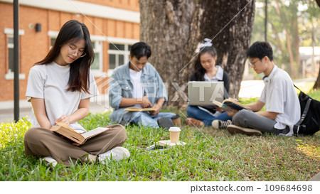 A focused Asian female student is reading a book while relaxing in a campus park with her friends. 109684698