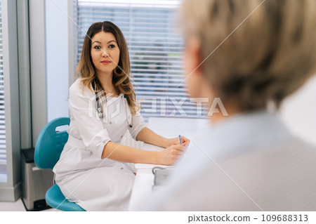 Young female doctor in white coat listening of unrecognizable senior aged woman patient tell about chronic disease, health complaints, affliction symptoms. Therapist take notes, fill history form. Young female doctor in white coat listening of unrecognizable senior aged woman patient tell about chronic disease, health complaints, affliction symptoms. Therapist take notes, fill history form. 109688313