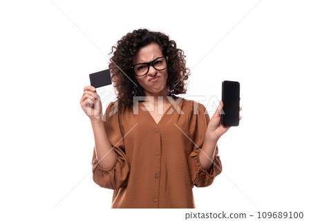 young curly businesswoman dressed in a brown shirt holds a phone and a card with a mockup on a white young curly businesswoman dressed in a brown shirt holds a phone and a card with a mockup on a white 109689100