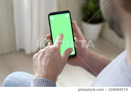 Young man sitting at home holding smartphone green mock-up screen in hand. Male person using chroma key mobile phone. Vertical mode. Touching, swiping display, tapping, surfing Internet social media Young man sitting at home holding smartphone green mock-up screen in hand. Male person using chroma key mobile phone. Vertical mode. Touching, swiping display, tapping, surfing Internet social media 109689574